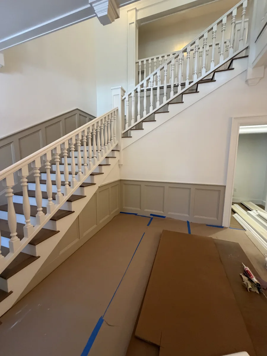 Staircase with white-painted balusters and greige wainscot panelling, floors covered during finishing